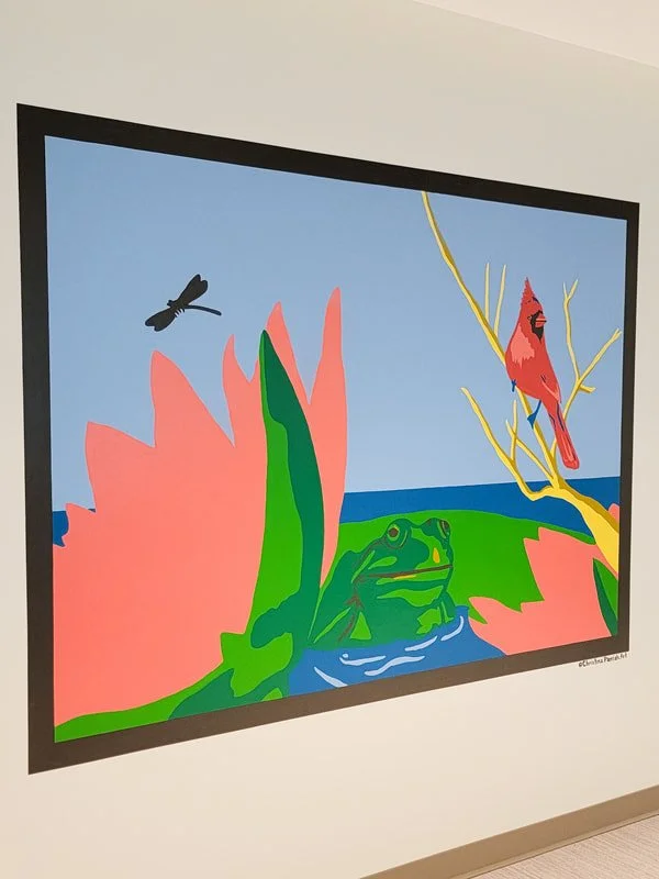 Colorful artwork featuring a frog partially hidden among large pink lily pads, a red cardinal bird perched on a yellow branch, a flying dragonfly, the sky and water in the background.