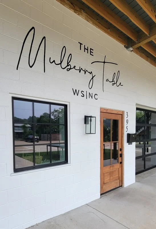 Exterior of a building with a white brick wall, a wooden door, and large windows. Handwritten-style sign on the wall reads 'The Mulberry Table WS|NC'.