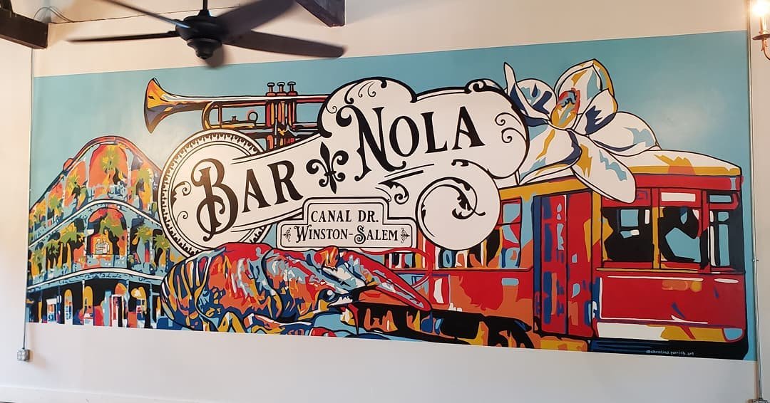 Colorful mural of NOLA Bar on Canal Drive, Winston-Salem, with a train, a trumpet, a fleur-de-lis, a flower, and a roller coaster.