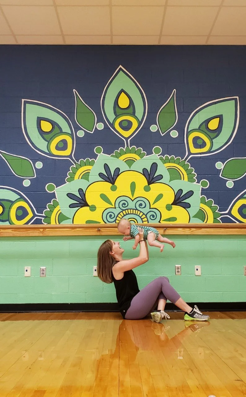 A woman sitting on the floor in a gym, lifting a baby above her with both hands. The woman has shoulder-length brown hair and is wearing a black tank top and purple leggings. The baby is wearing a striped onesie and is smiling while looking down at t