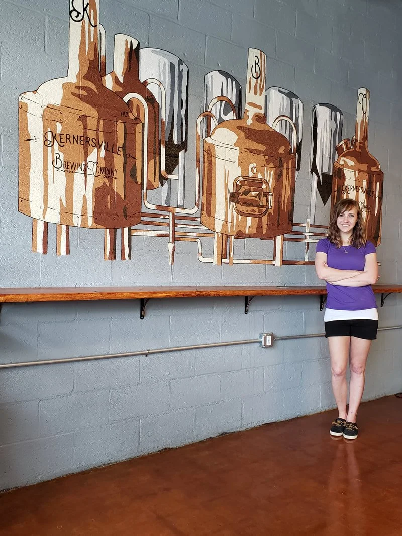 A young woman in a purple shirt and black shorts stands with crossed arms and smiling in front of a mural of brewing equipment on a gray wall, inside a brewery or similar establishment.