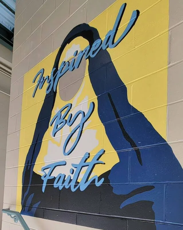 A mural of a woman with her hair up, made with blue, white, yellow, and black paint, displaying the words 'Inspired By Faith' in cursive script.