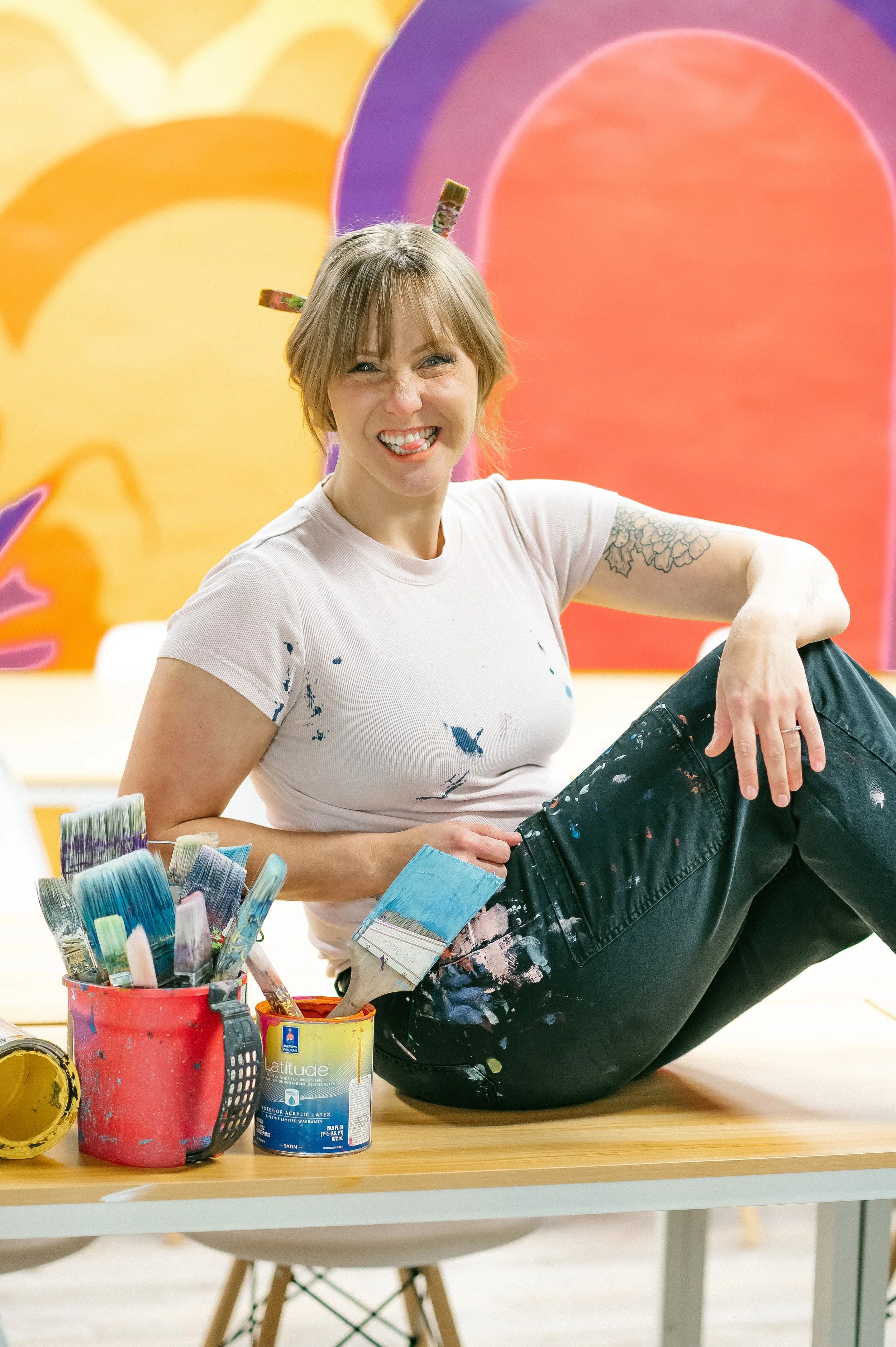 Woman with light brown hair, wearing a white t-shirt with paint splatters, sitting on the floor in front of a table with paint cans and brushes, smiling playfully with tongue out, colorful abstract background.