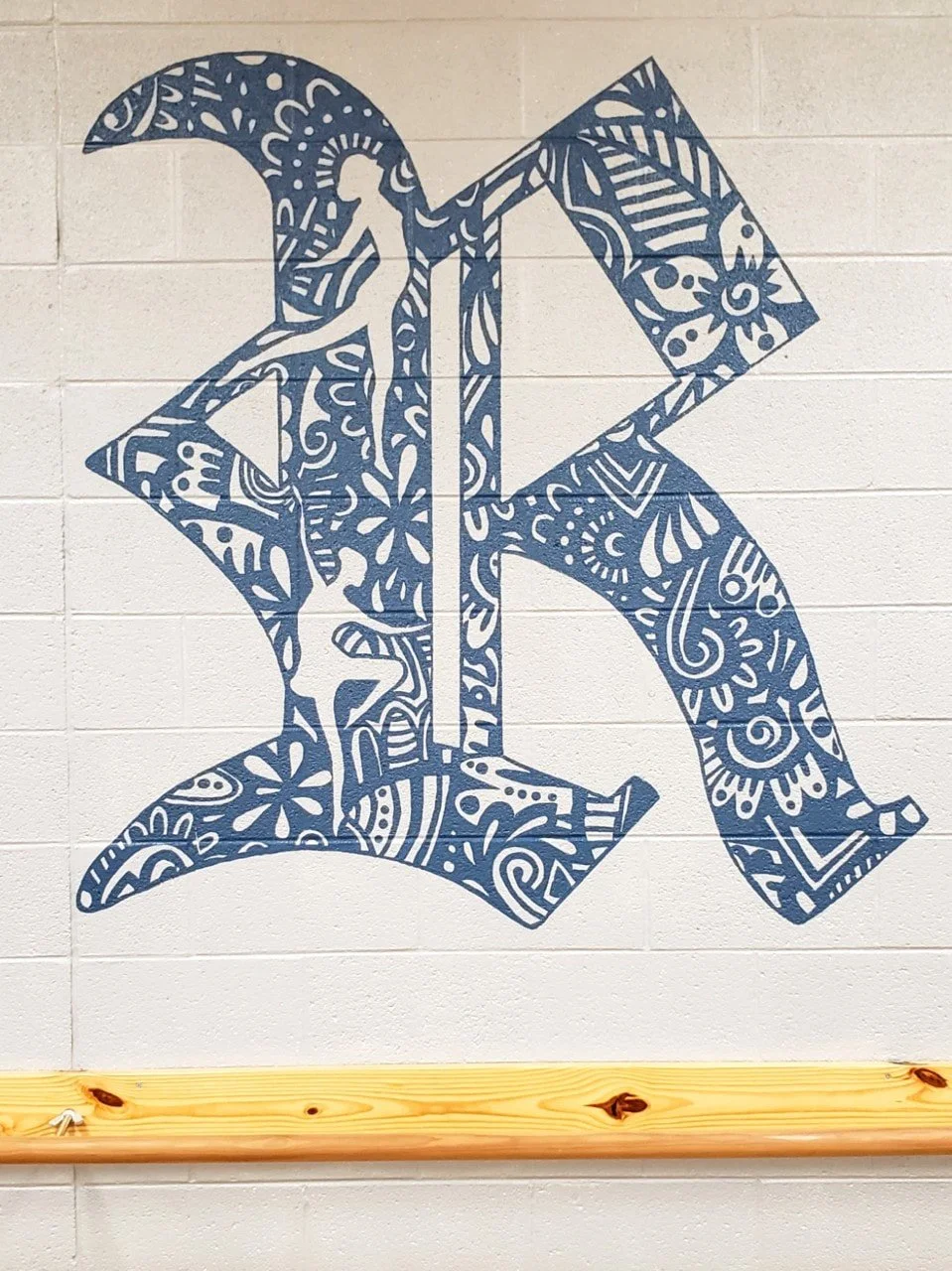 Decorative blue letter 'K' with intricate patterns and a small silhouette of a person sitting on the top left of the letter, mounted on a light-colored brick wall with a wooden rail at the bottom.