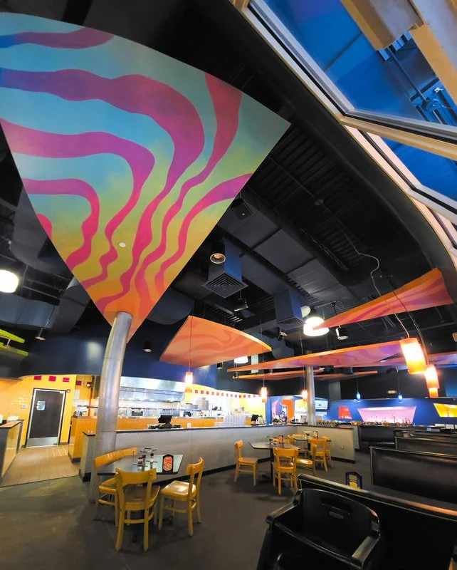 Interior of a modern restaurant with colorful geometric ceiling art, wooden chairs, and a bar area in the background.