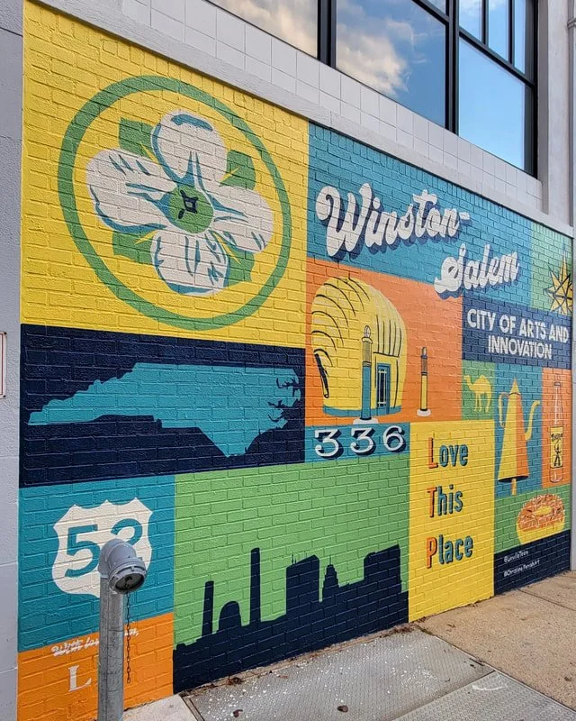Colorful mural on a brick wall featuring a flower, a map of North Carolina, a skyline, the numbers '336', the phrase 'Love This Place', and icons representing the city of Winston-Salem, NC.