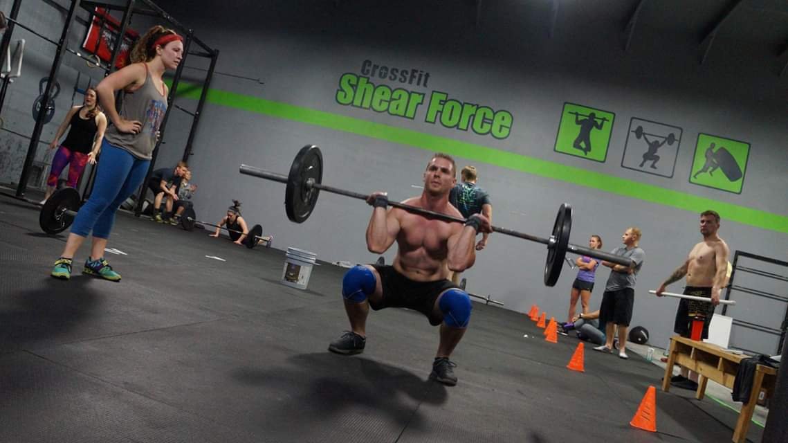 A muscular man performing a squat with a barbell at CrossFit Shear Force gym, surrounded by other people working out.