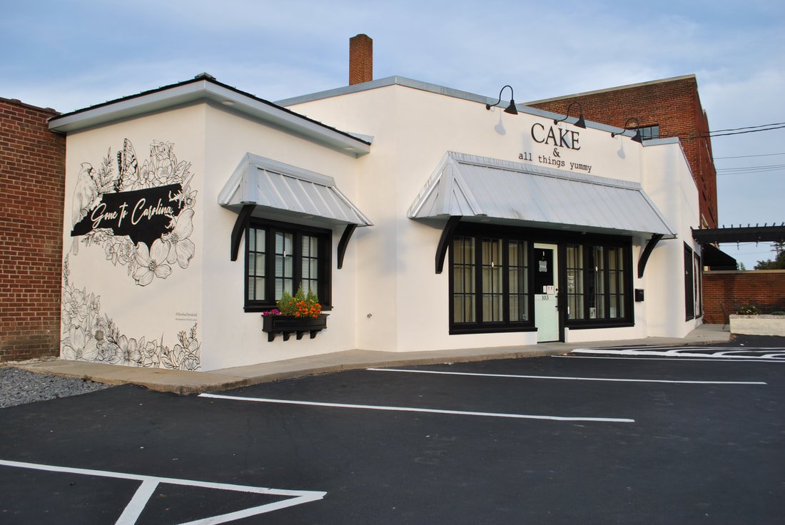 Front view of a small bakery with white walls, black window frames, and a sign that reads "Cake & all things yummy." A mural on the side wall features floral illustrations and the words "Grove & Carolina." The parking lot has marked spaces in front.