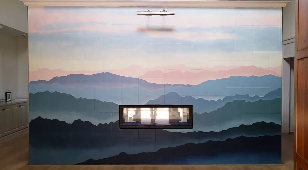 Wall mural of layered mountains at sunrise or sunset in pastel pink, blue, and green tones with a fireplace cutout in the center.