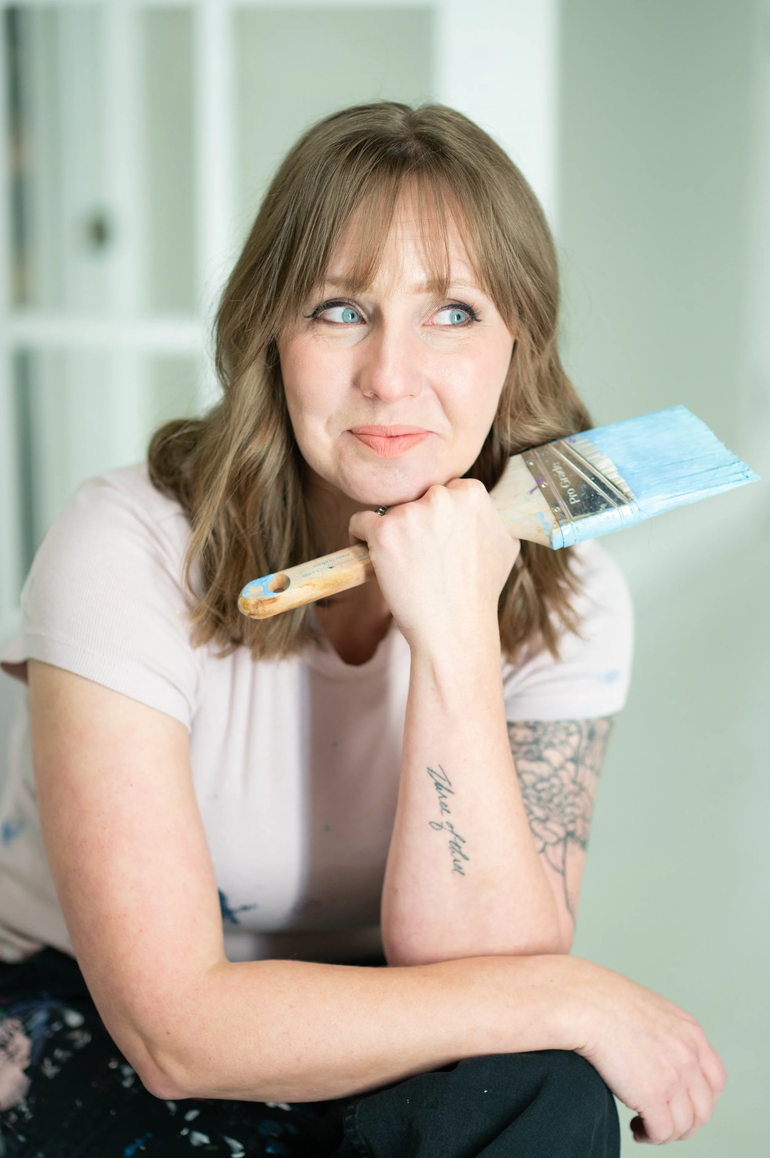 A woman with light brown hair and blue eyes, resting her chin on her hand, holding a paintbrush. She has tattoos on her arm and is wearing a light-colored shirt, sitting in a room with light green walls.