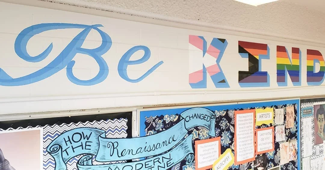 School bulletin board with colorful letters spelling "Be KIND" and various informational posters.