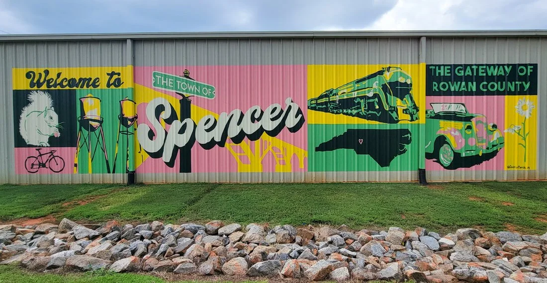 Colorful mural welcoming visitors to Spencer, a town in Rowan County. The mural features a cat, two chairs on tall legs, a bicycle, a train, a vintage car, and flowers, with the text 'Welcome to The Town of Spencer' and 'The Gateway of Rowan County.'