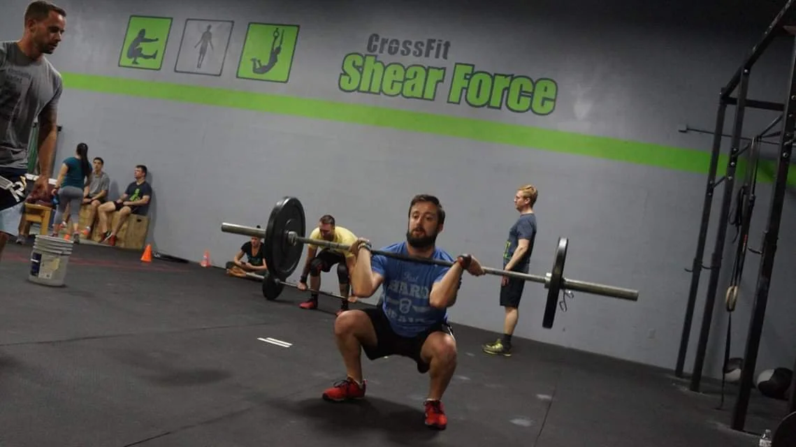 A man is performing a squat lift with a barbell at CrossFit Shear Force gym, with others in the background and gym equipment visible.