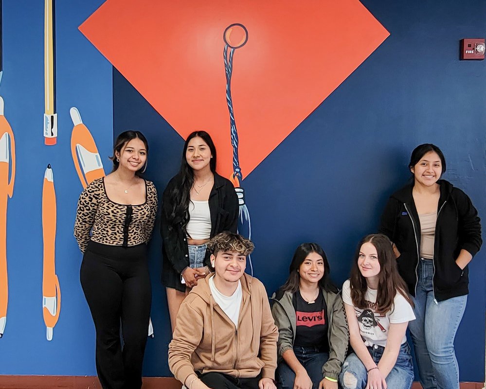 Group of six young people posing in front of a blue wall with orange painted art, including a large hanged loop and pencil illustrations.