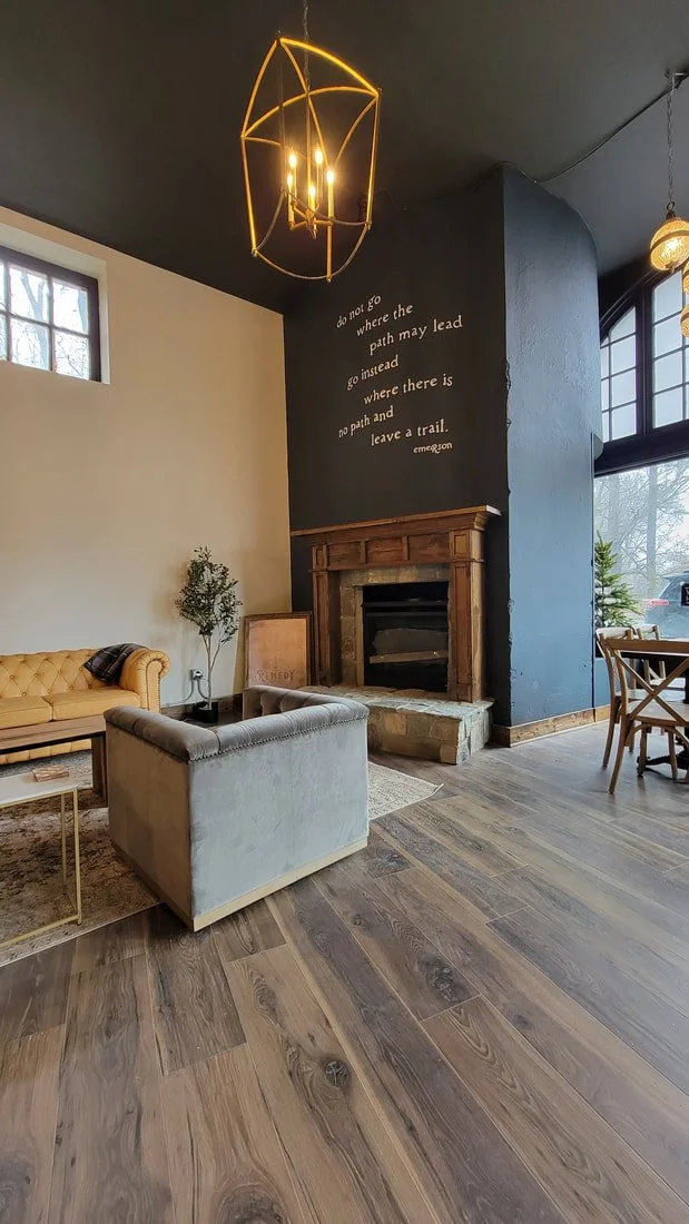 Living room with a dark blue accent wall, a wooden fireplace mantel, a beige tufted sofa, a gray velvet armchair, a small plant, a framed picture, a rug, a modern chandelier, and large windows letting in natural light.