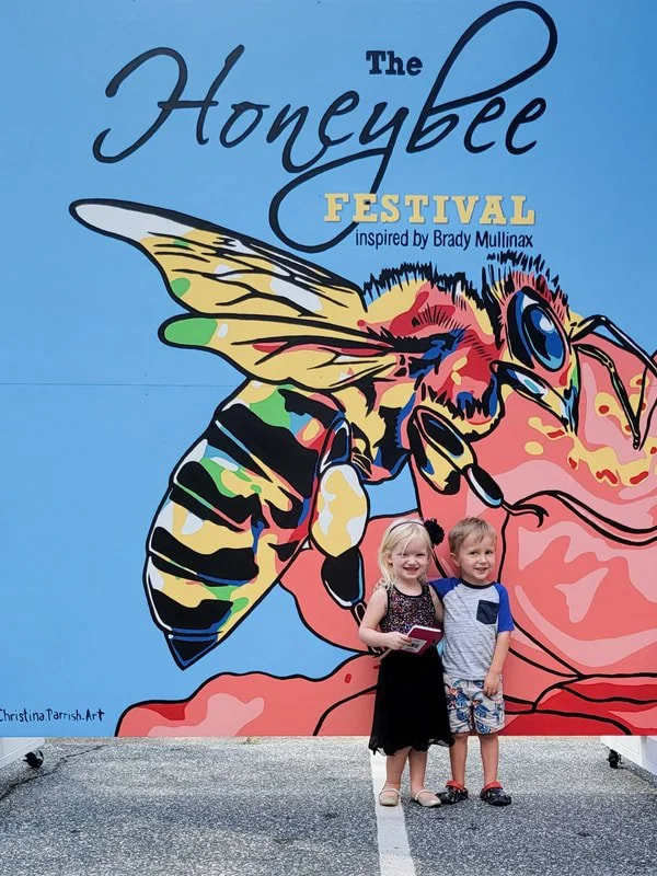 Two children, a girl and a boy, stand in front of a colorful mural for The Honeybee Festival inspired by Brady Mullinax, featuring a large, stylized bee and a smiling woman. The girl holds a book and the boy has his arm around her, both smiling.