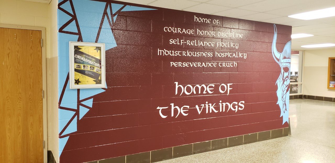 School hallway mural with a Viking helmet illustration on the right and words on the wall that read "home of the vikings." The wall is painted in red and light blue, with some geometric designs and a framed poster on the left.