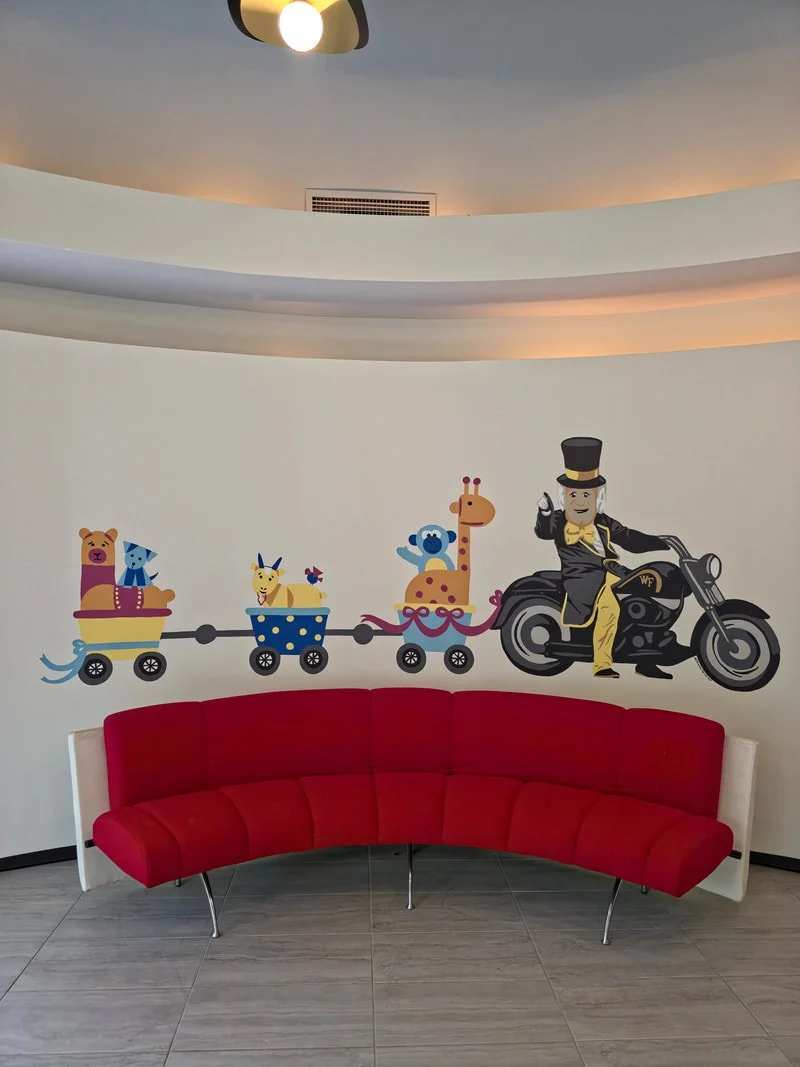 Colorful cartoon wall mural of a train with animal characters, including a bear, dog, giraffe, and bird, being driven by a person with a top hat on a motorcycle, in a modern room with a red curved sofa and a ceiling light.