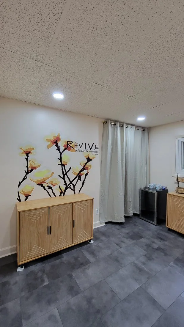 Interior room with a wall mural of a flowering branch, a wooden cabinet, a small black refrigerator, a wicker basket, a window with closed curtains, and white cups in a stack.