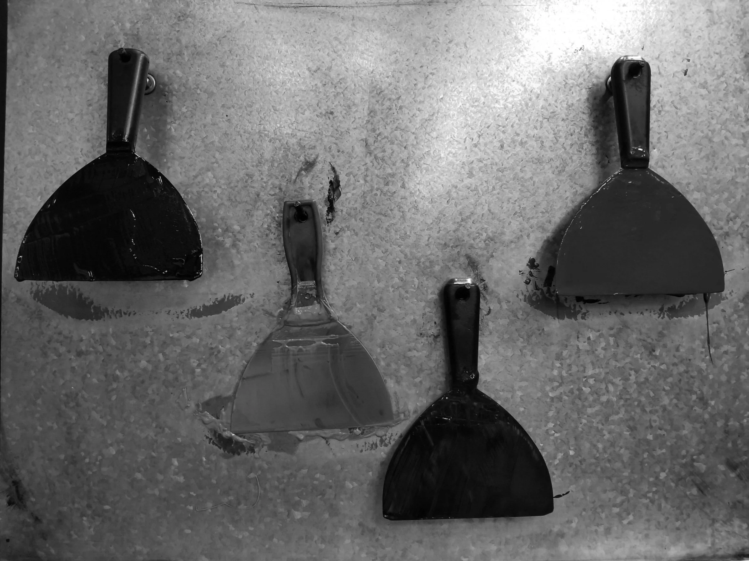 Four wall scrapers with varied blade sizes hanging on a textured metal surface in black and white.