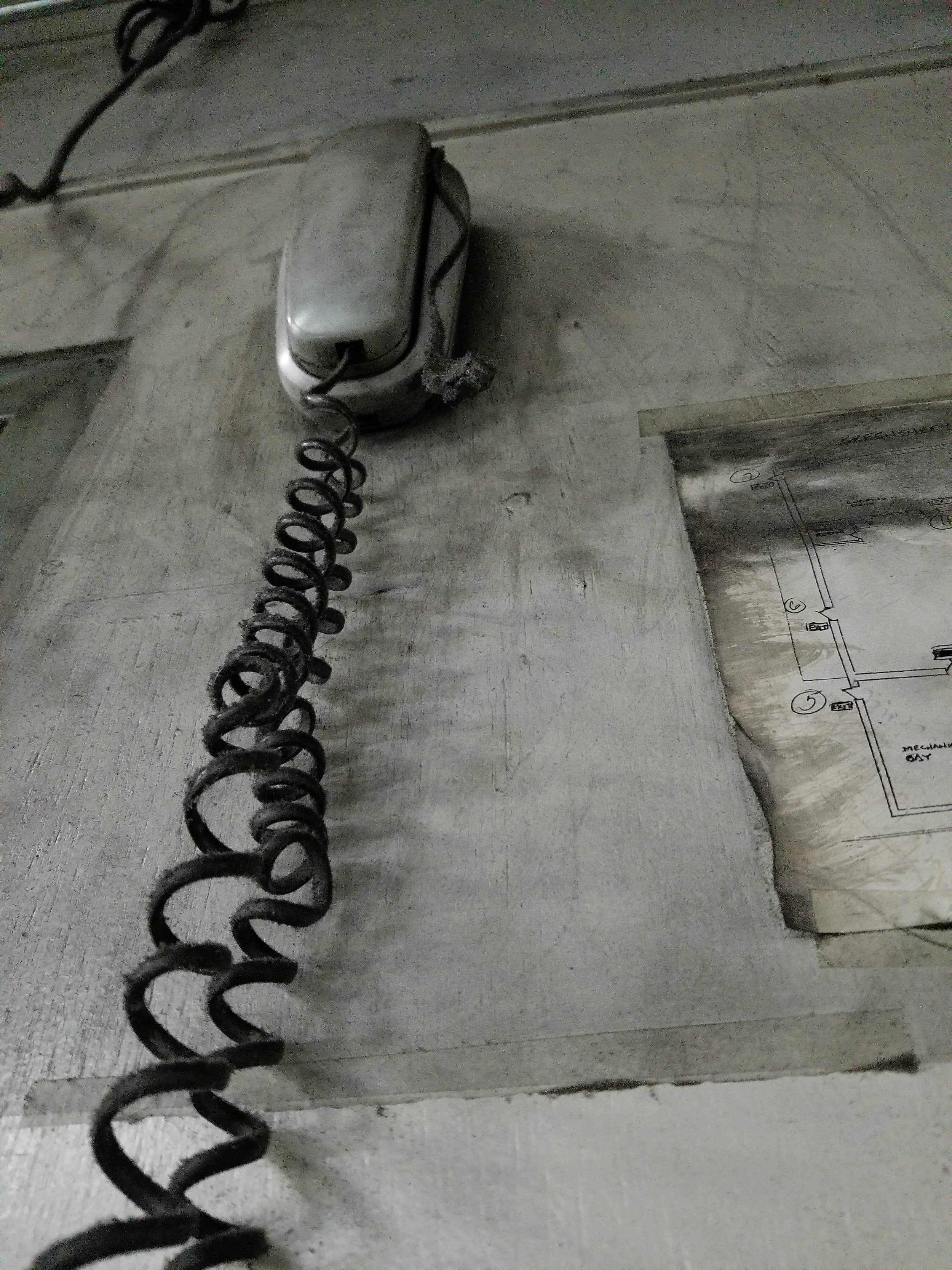 Old metallic wall-mounted telephone with coiled cord and a faded paper diagram on a textured surface.