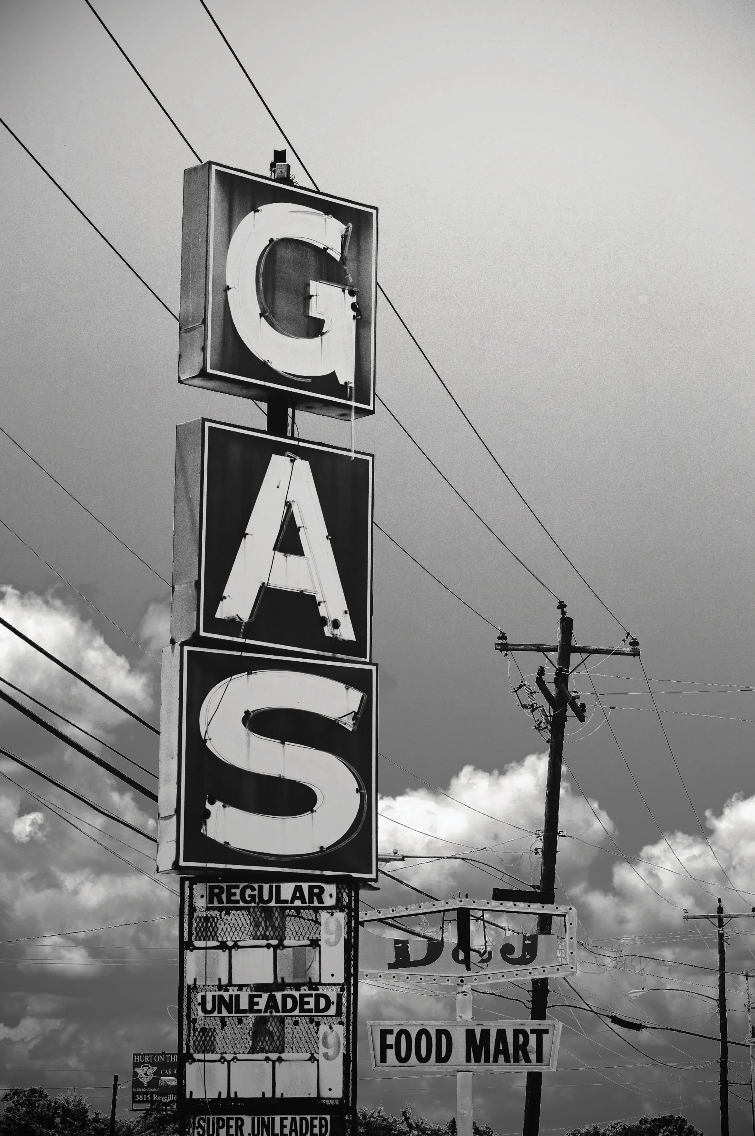 Black and white photo of vintage gas station sign reading 'GAS' with additional signs for 'Regular Unleaded' prices and 'Food Mart,' accompanied by power lines against a cloudy sky.