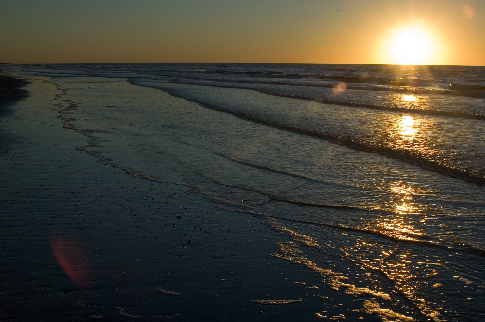 Sunset at the beach with the sun reflecting on the water and gentle waves coming ashore.