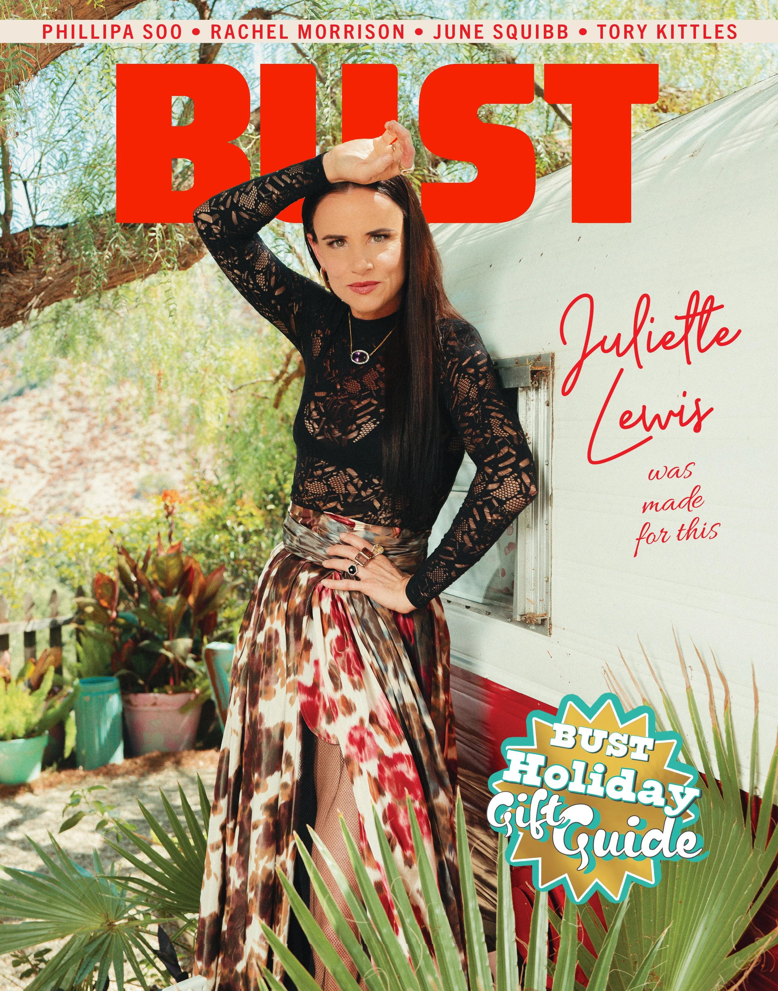 Cover of Bust magazine featuring Juliette Lewis in a black lace top and multicolored skirt, standing outdoors with greenery, plants, and a white trailer. Large red text reads 'BUST'; smaller text above lists names. Handwritten-style red text says 'Juliette Lewis was made for this.' A badge in the bottom right corner reads 'Bust Holiday Gift Guide.'