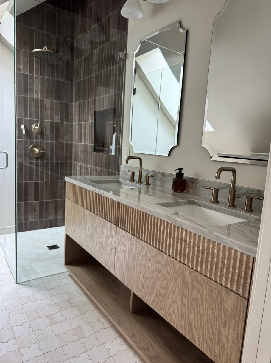 Modern bathroom with a double vanity, marble countertop, brass fixtures, and a floor-to-ceiling tiled shower area with dark brown tiles and a glass door.