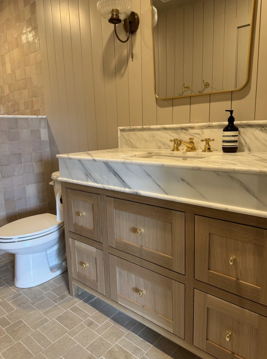 A bathroom featuring a wooden vanity with a marble countertop and gold hardware, a white toilet, a wall mirror with a gold frame, a wall-mounted light fixture, and a soap dispenser.