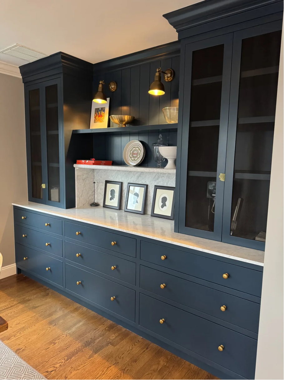 A navy blue built-in cabinet with glass doors, gold knobs, and a white marble countertop, decorated with framed silhouette photographs, decorative bowls, and tray, with two gold and black wall sconces and a small colorful painting above.