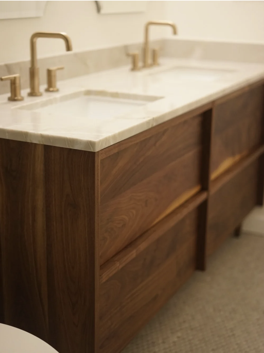 A bathroom vanity with a white marble countertop, gold faucet fixtures, and walnut cabinet drawers.
