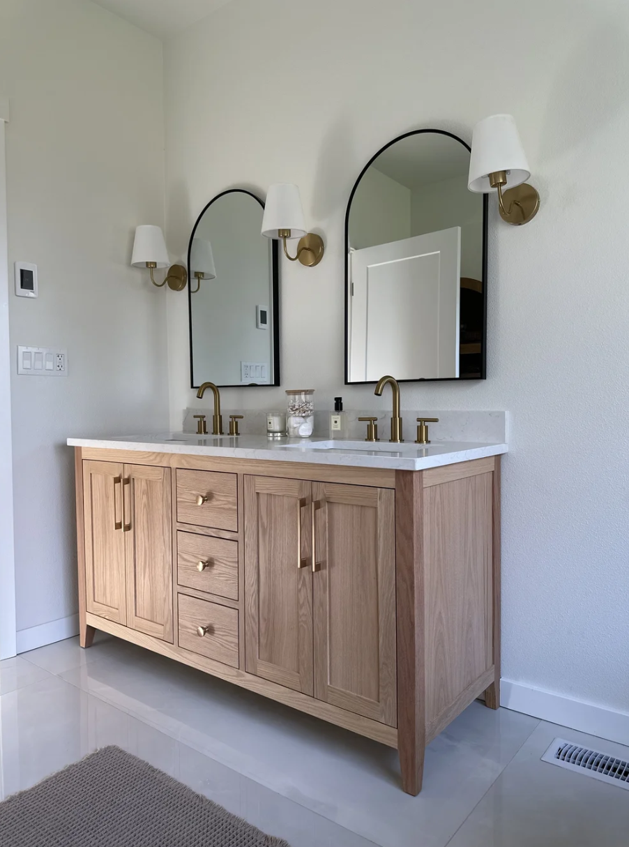 A modern bathroom vanity with a light wood cabinet, white marble countertop, two brass faucets, and two arched mirrors. Wall-mounted light fixtures with white shades next to each mirror.