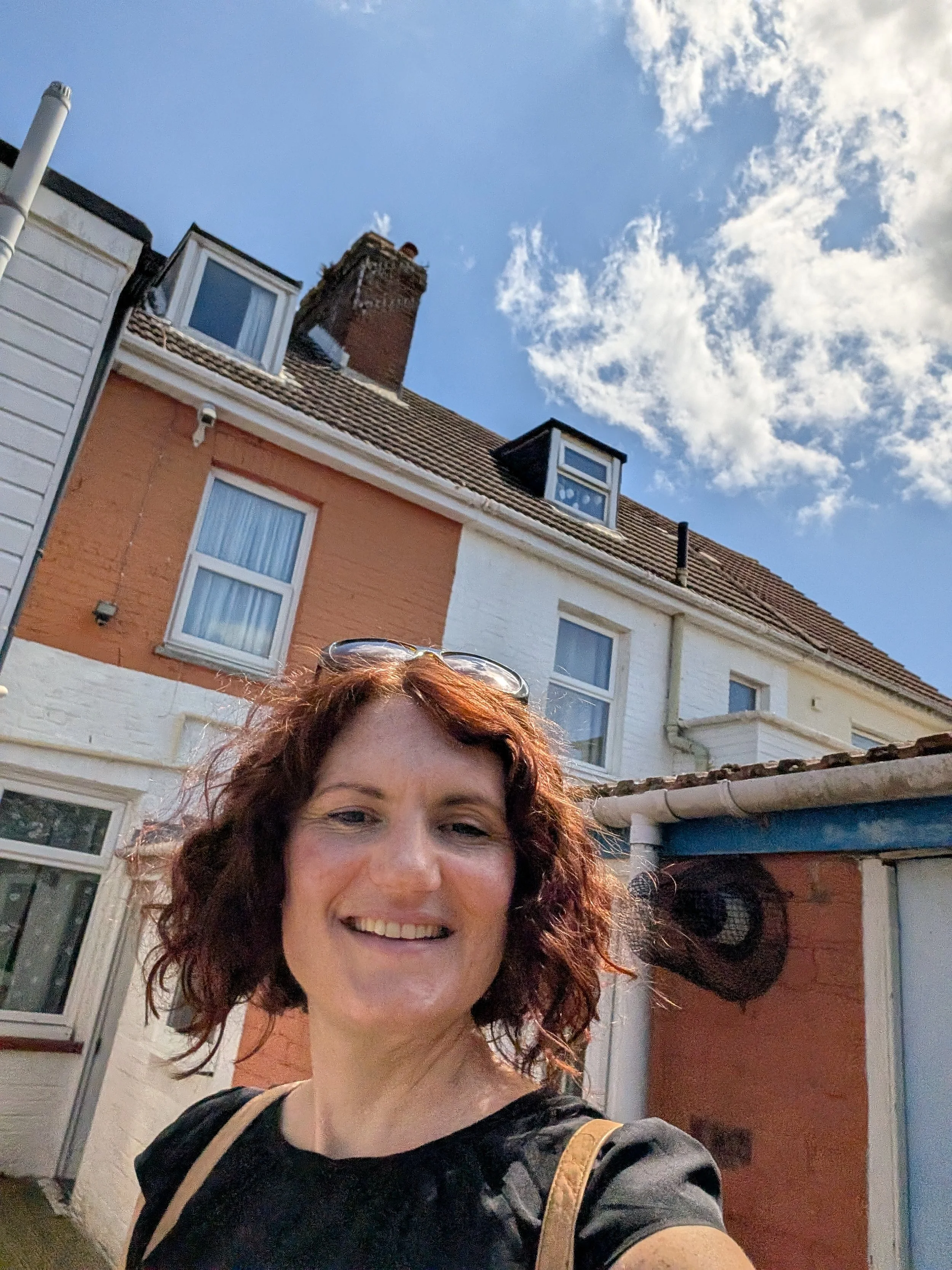 A woman with curly red hair smiling and taking a selfie outside in front of houses with brick walls, windows, and a chimney under a blue sky with some clouds.