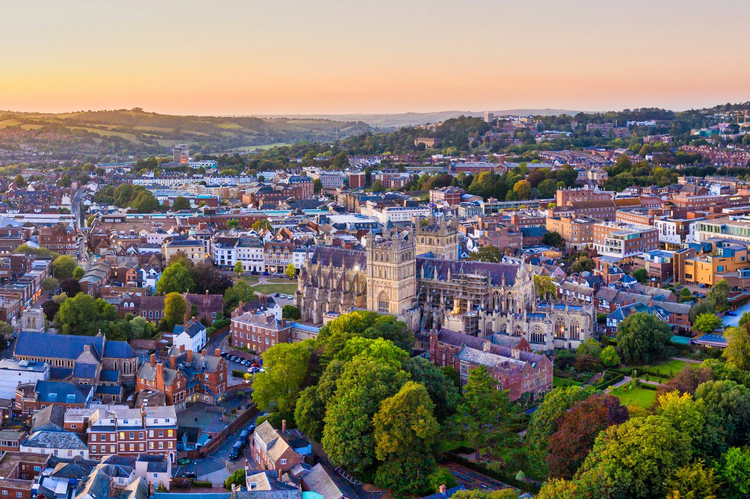 Aerial view of a cityscape with a historic Gothic cathedral, modern buildings, and rolling green hills in the background during sunset.