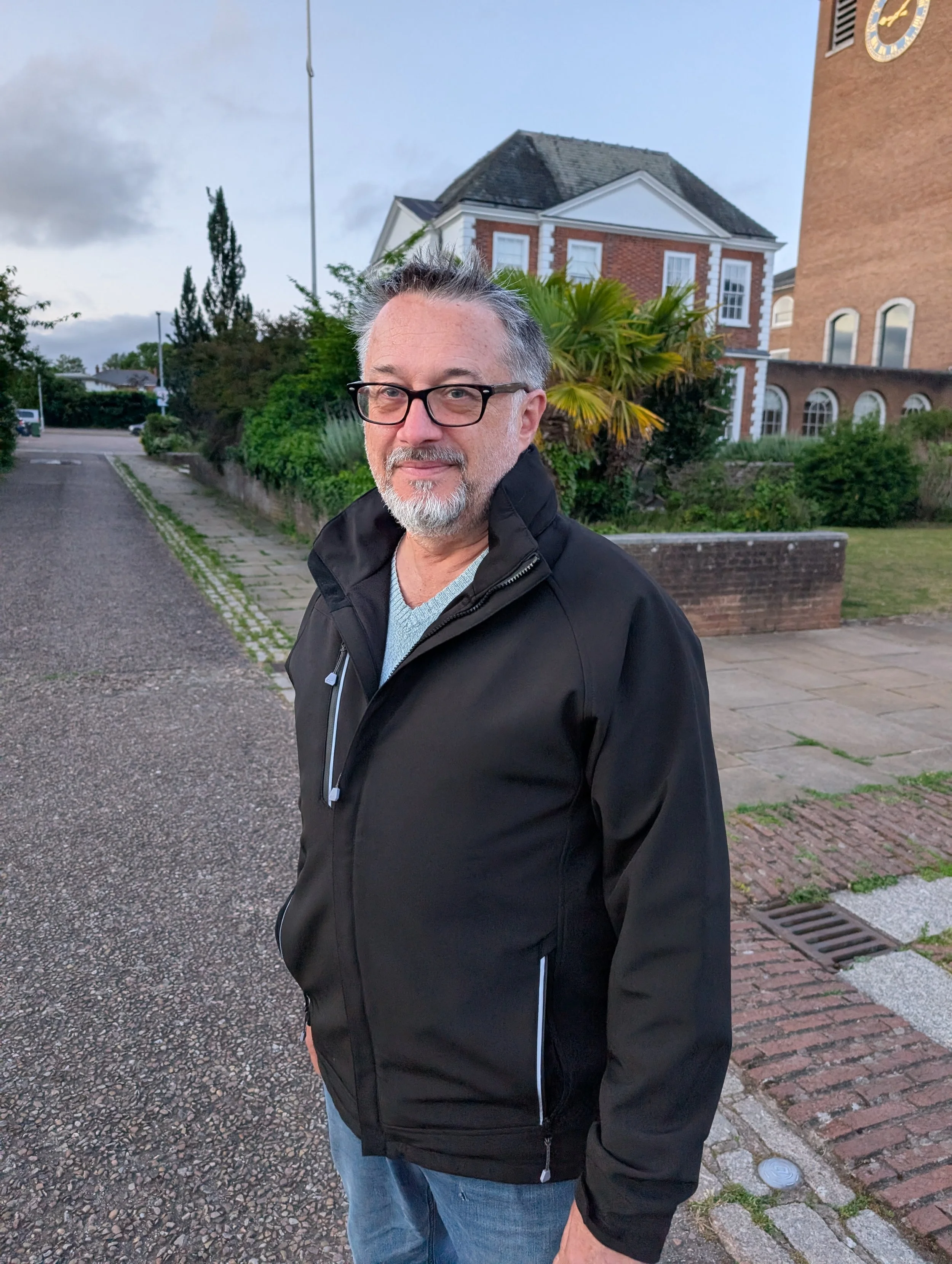 A middle-aged man with glasses, gray beard, black jacket, and blue jeans standing outdoors near a sidewalk, with a brick building and greenery in the background.