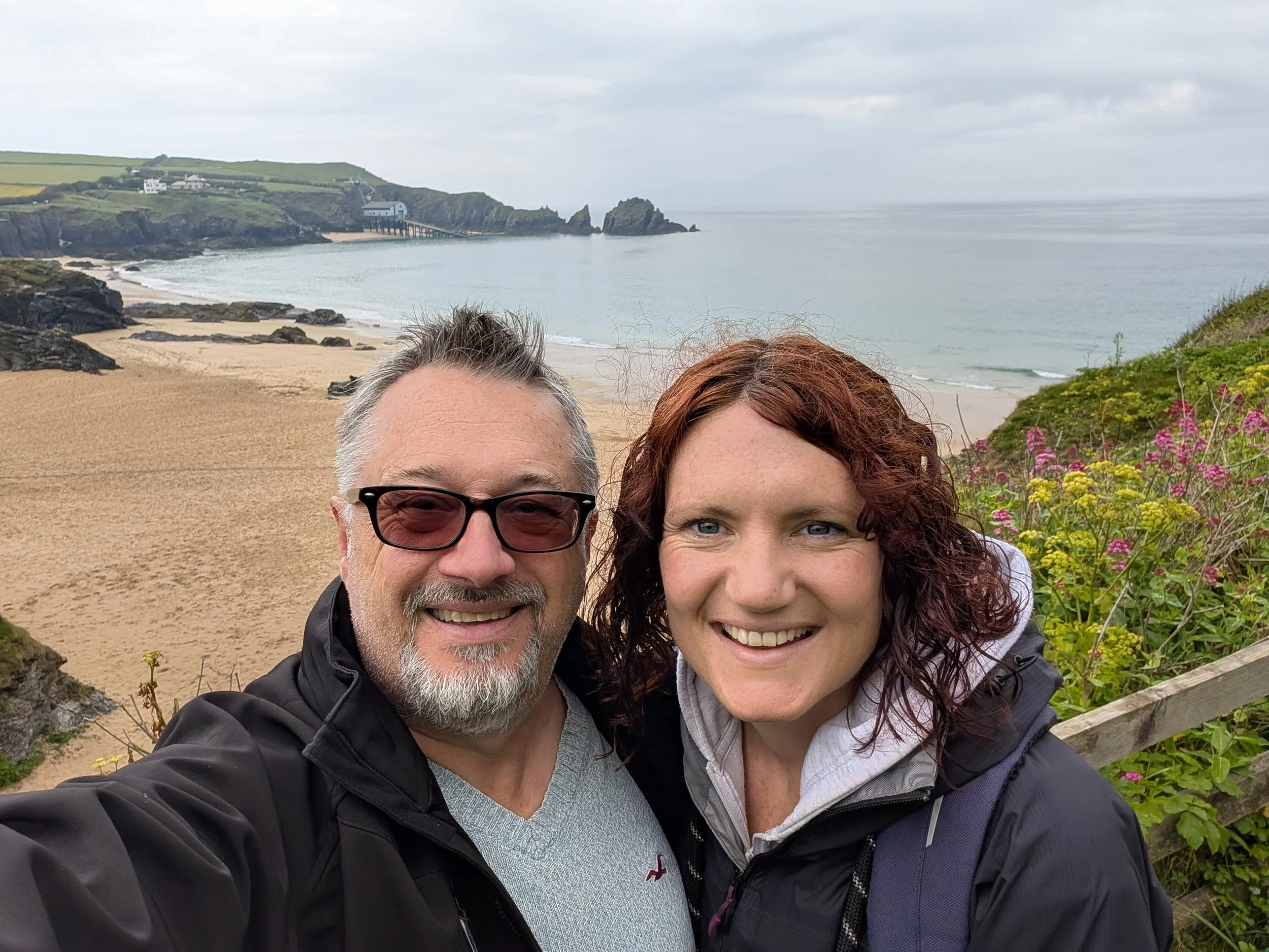 A smiling older man with glasses and a beard taking a selfie with a woman with curly red hair at a beach with cliffs, rocks, sand, and a pier in the background.