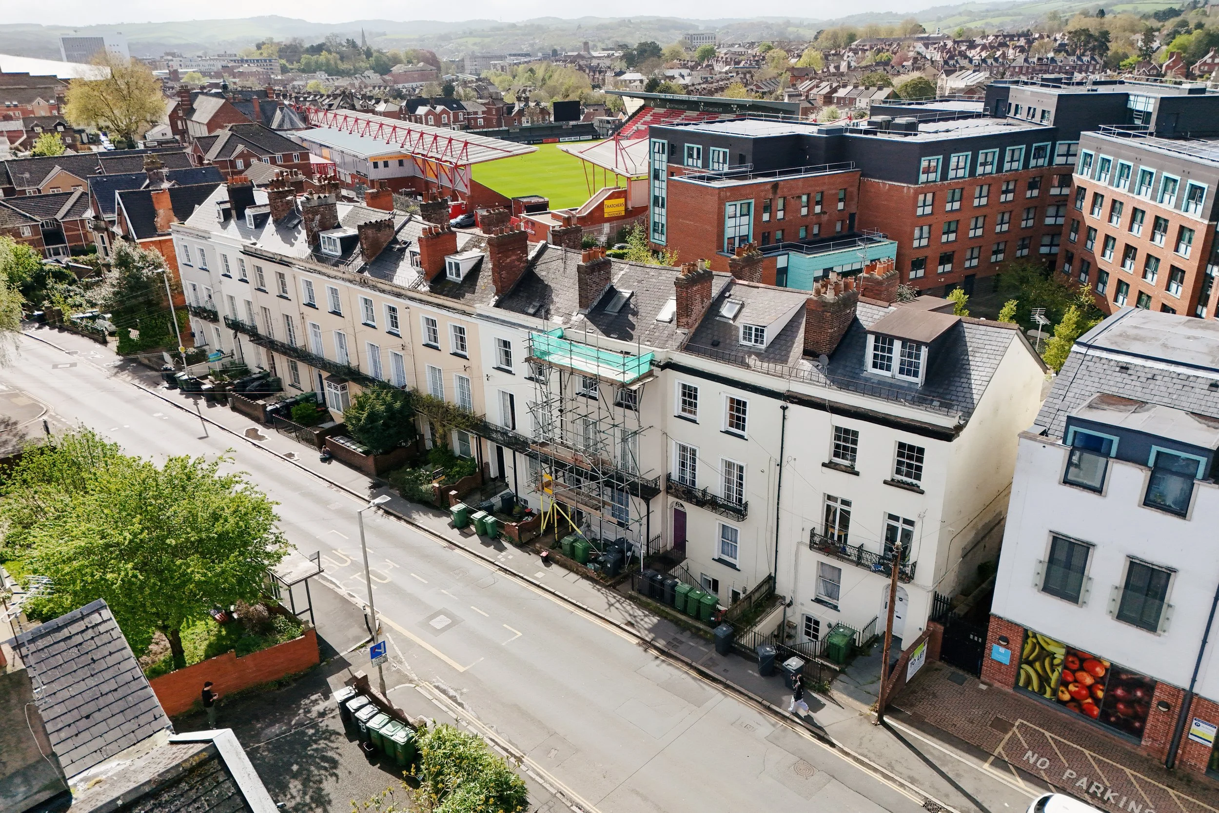 Aerial view of a cityscape featuring a rugby stadium with red seats, streets lined with residential houses, and a mix of modern and traditional buildings amidst green hills in the background.