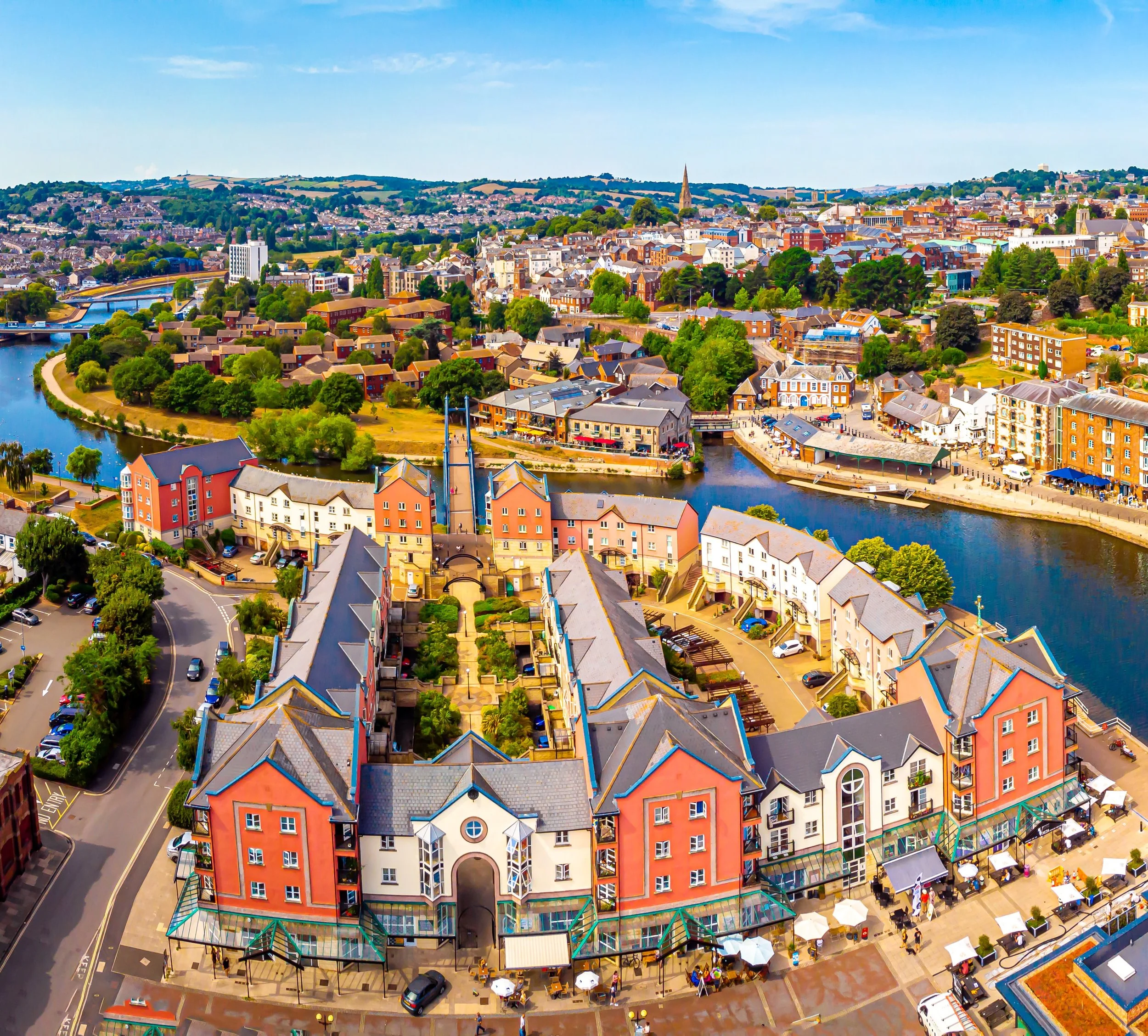 Aerial view of a colorful, residential seaside town with a river flowing through it, multiple bridges, boats, and buildings, featuring a prominent orange and white building with a pub-like appearance and outdoor seating.