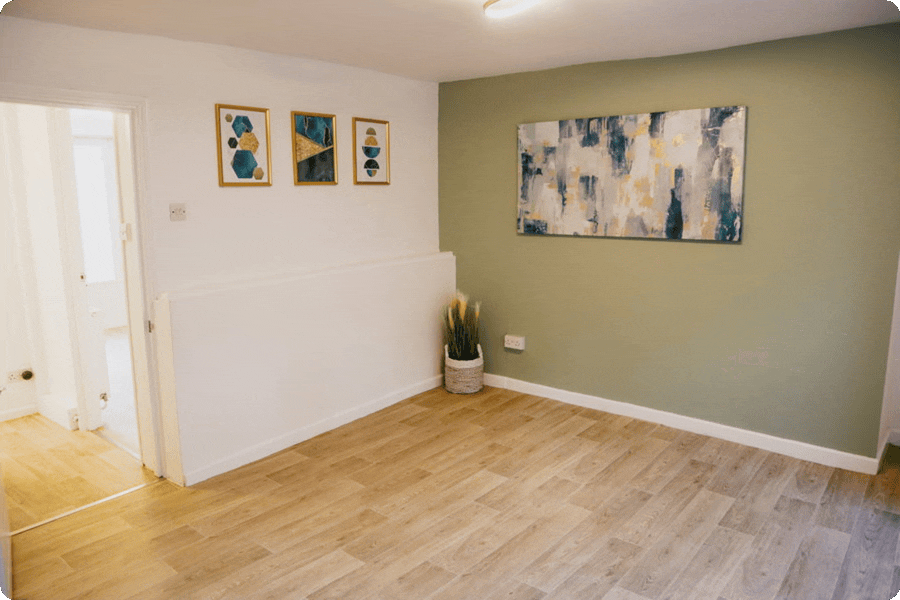 Empty living room with a green accent wall, wooden floor, modern abstract paintings, and a potted plant in a white basket.