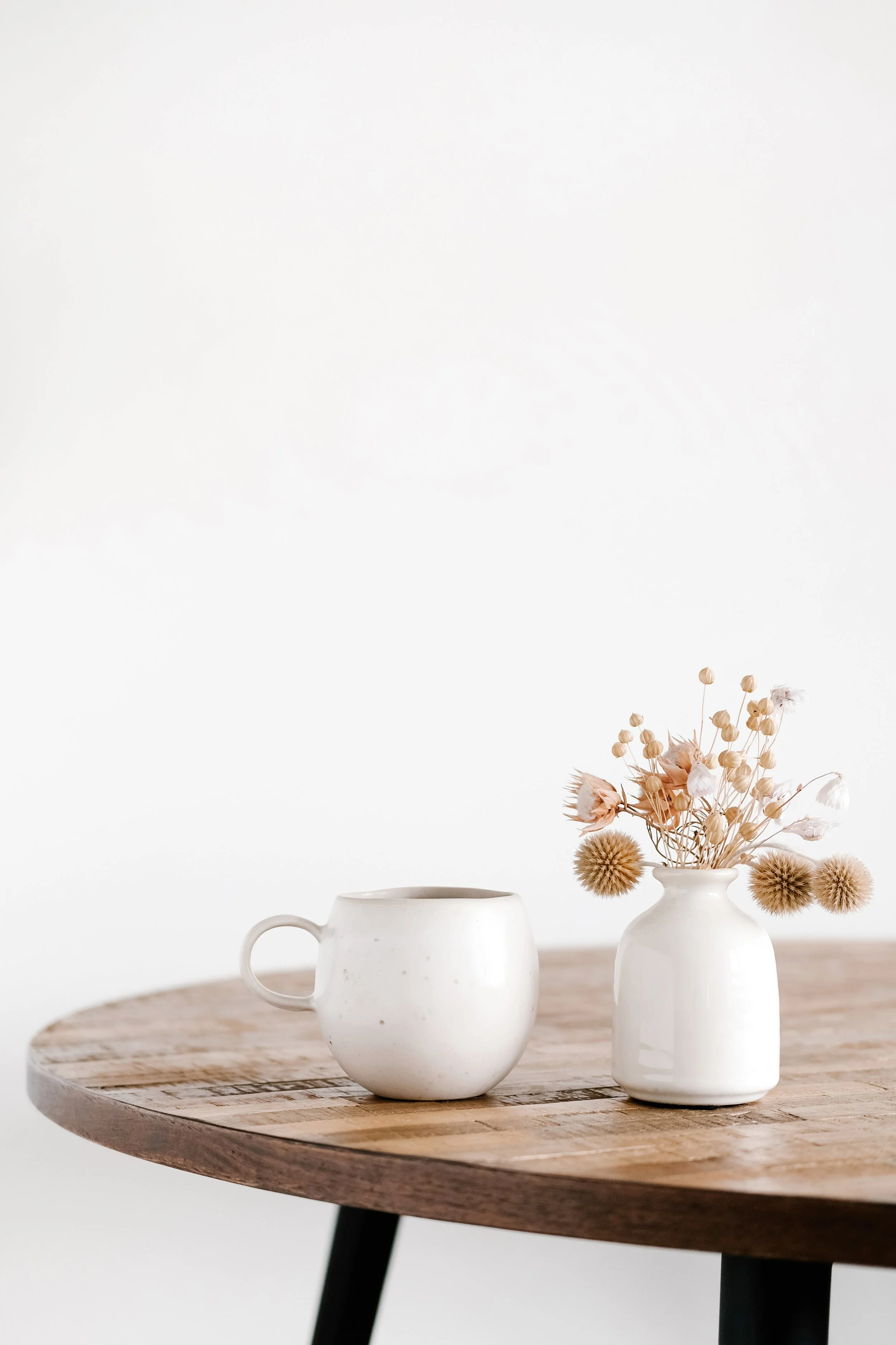 White ceramic mug and two white vases containing dried flowers on a wooden table against a plain white background.