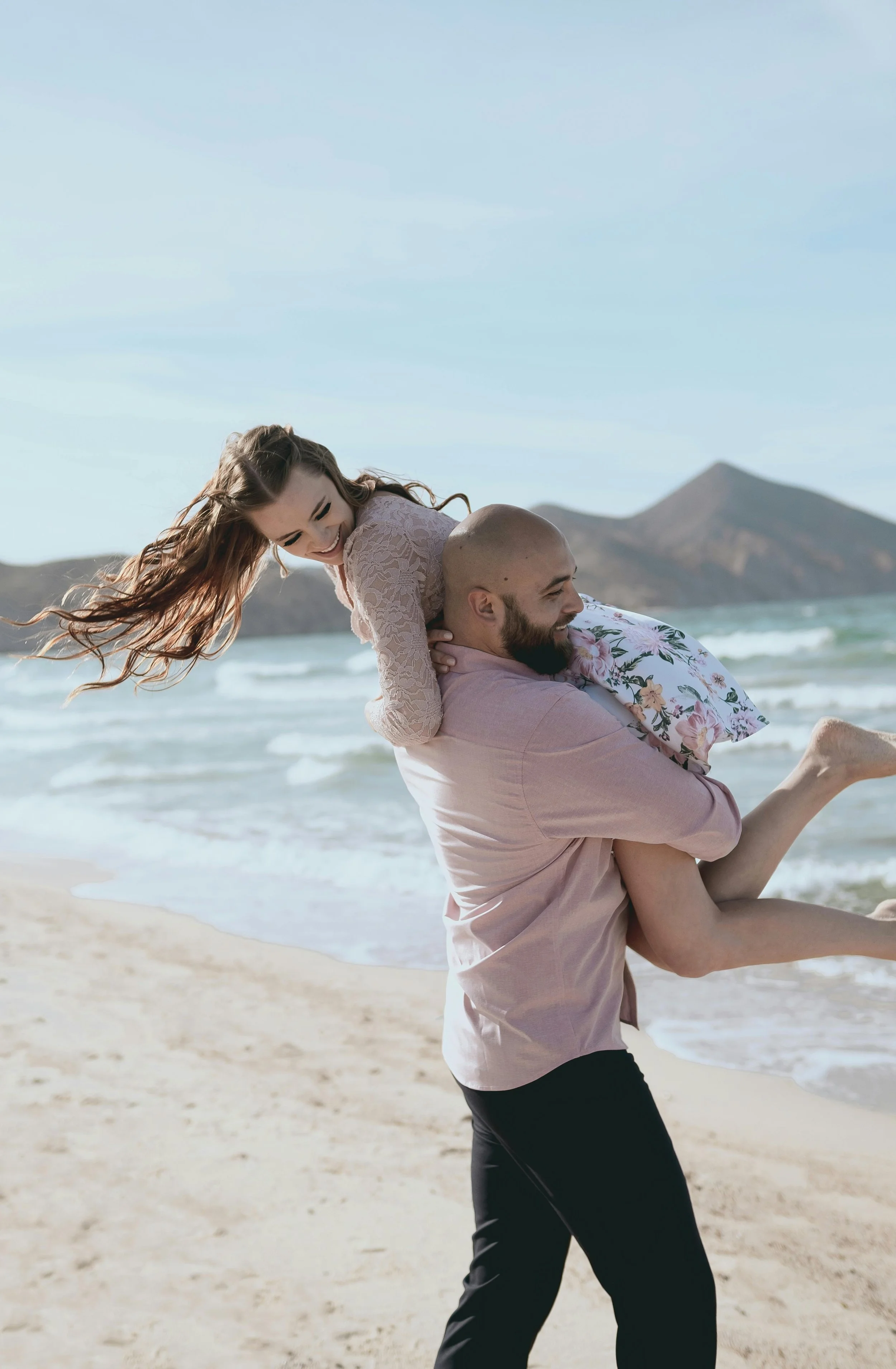 A man giving a woman a piggyback ride on a sandy beach with ocean waves and mountains in the background.