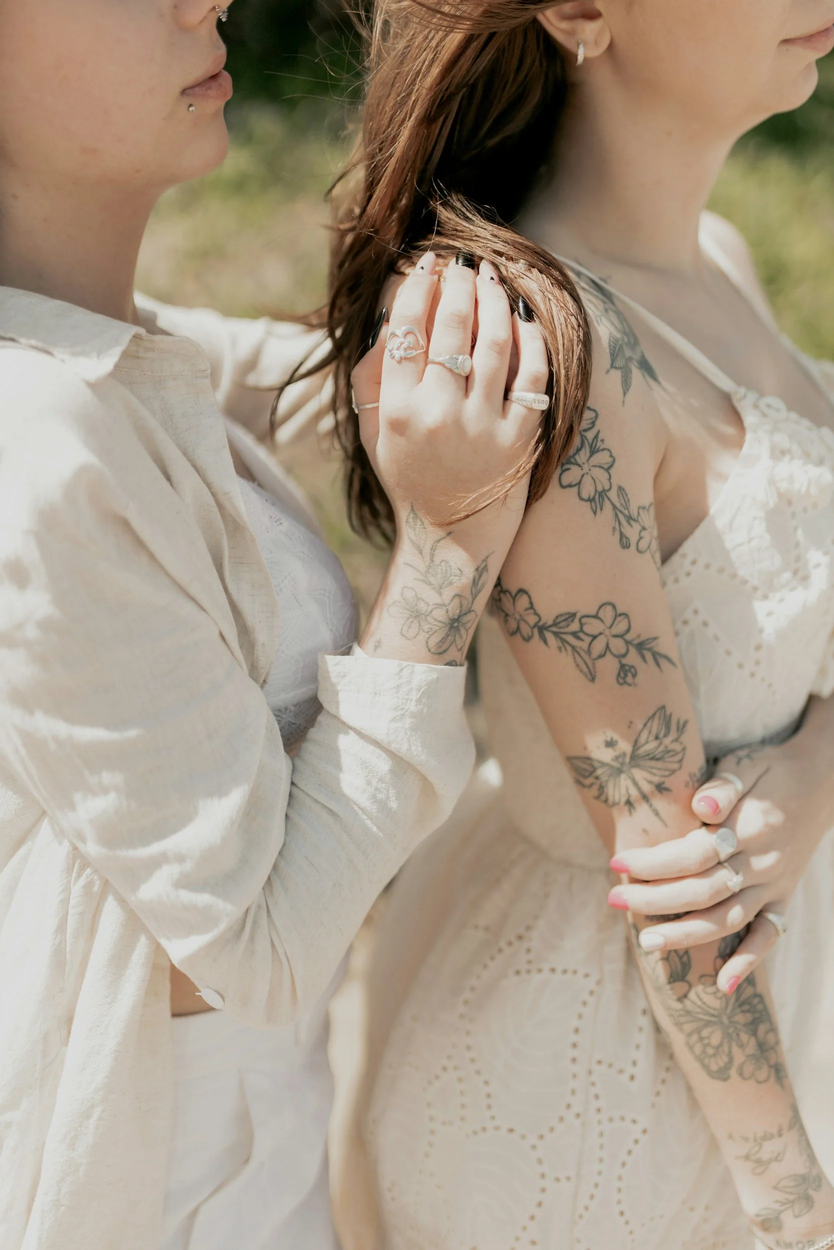 Two women are standing close together outdoors, both wearing light-colored dresses. The woman on the left has dark hair and is touching the shoulder of the woman on the right, who has tattoos on her arm and shoulder. They are both wearing rings and jewelry, and the woman on the left has dark nail polish. The background is blurred, suggesting a sunny day.