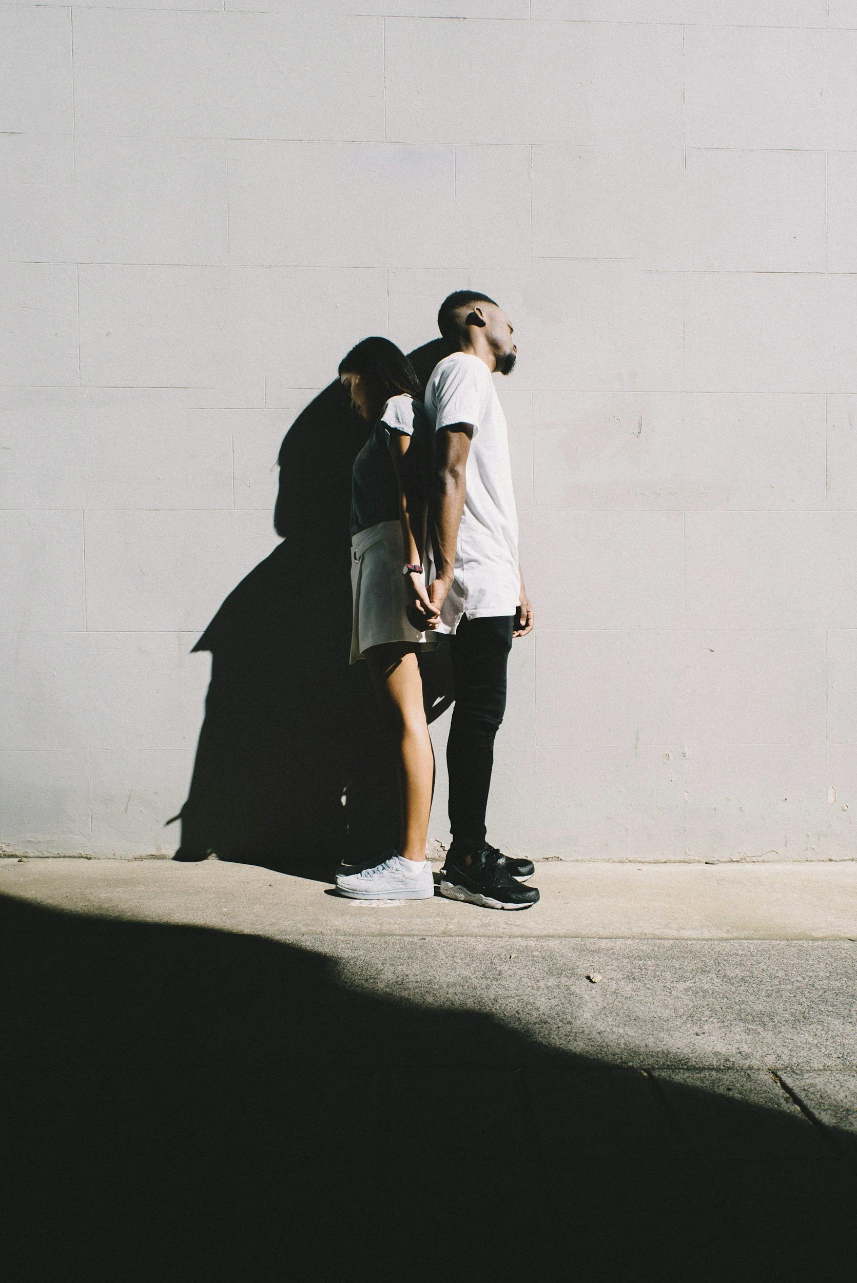 A man and a woman stand against a plain wall with their shoulders touching, with their shadows cast on the wall behind them. The man is wearing a white t-shirt, black pants, and black sneakers; the woman is wearing a black t-shirt, light-colored shorts, and white sneakers.
