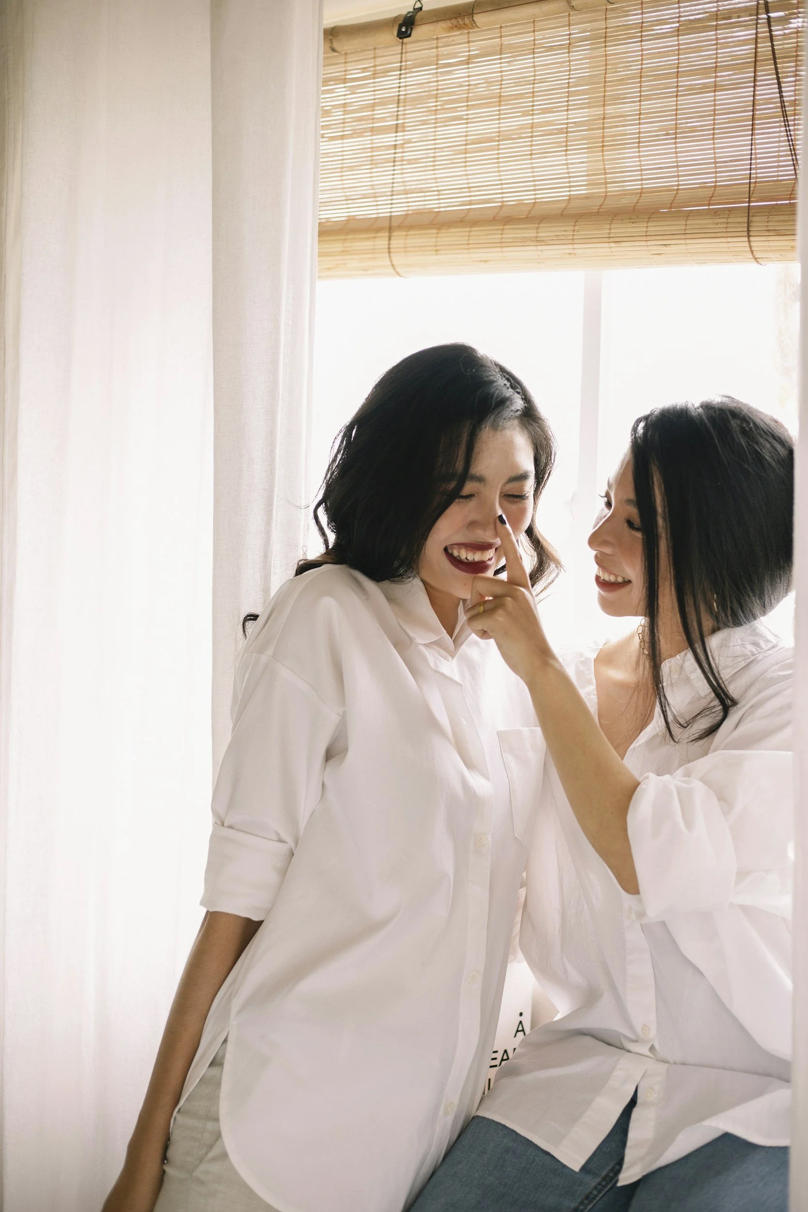Two women wearing white shirts sharing a joyful moment in front of light-colored curtains and a bamboo window blind, with sunlight illuminating the room.