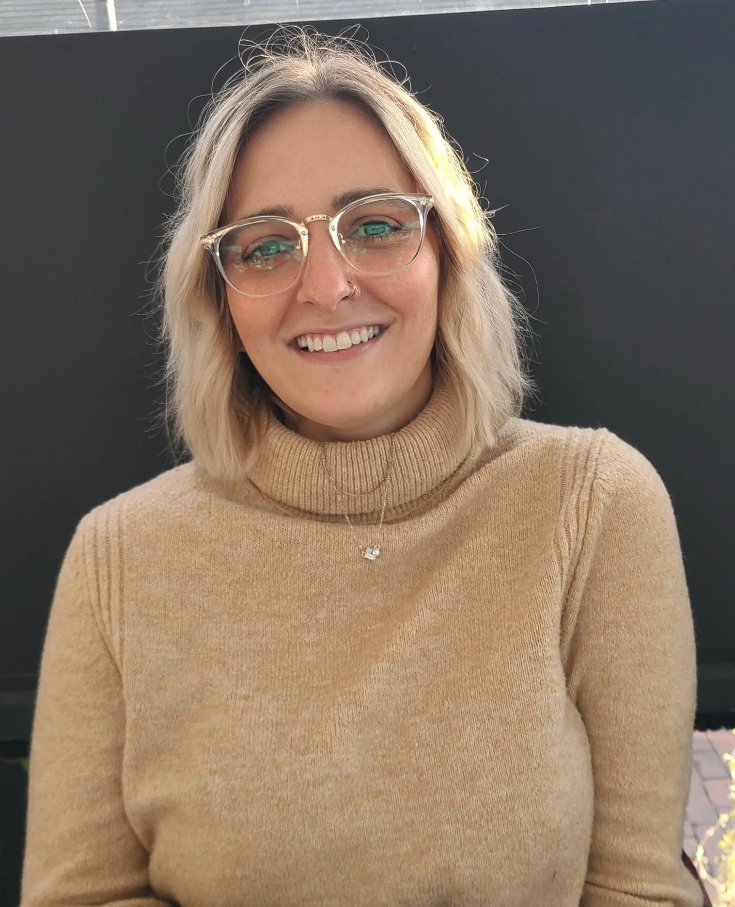 A woman with blonde hair, wearing glasses and a beige turtleneck sweater, smiling in front of a black background.