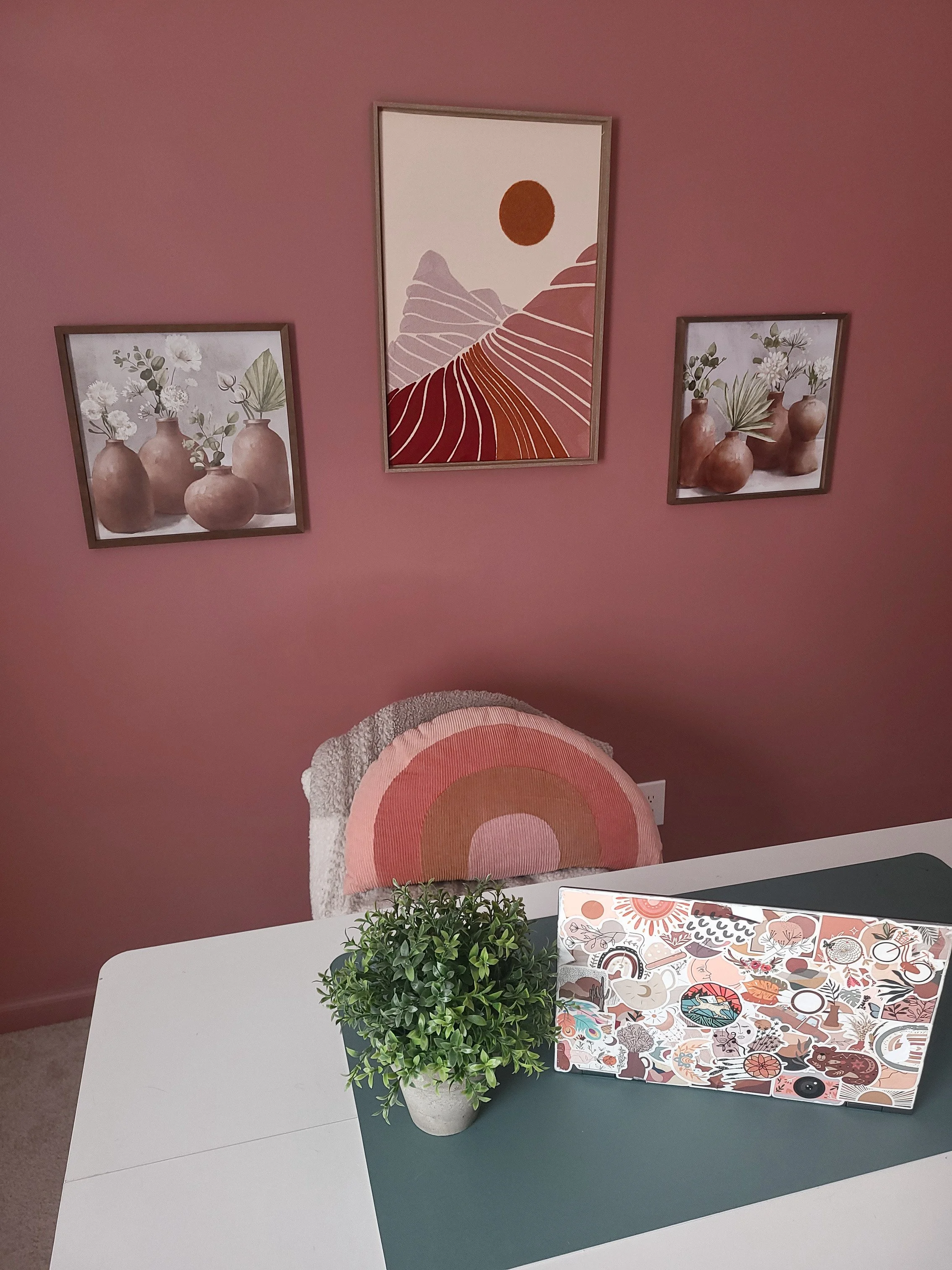 A white table with a potted plant, a colorful sticker sheet, and a patterned folder. Behind the table, there is a pink wall with three framed artworks: two with brown vases holding flowers, and one abstract landscape with a sun and mountains.
