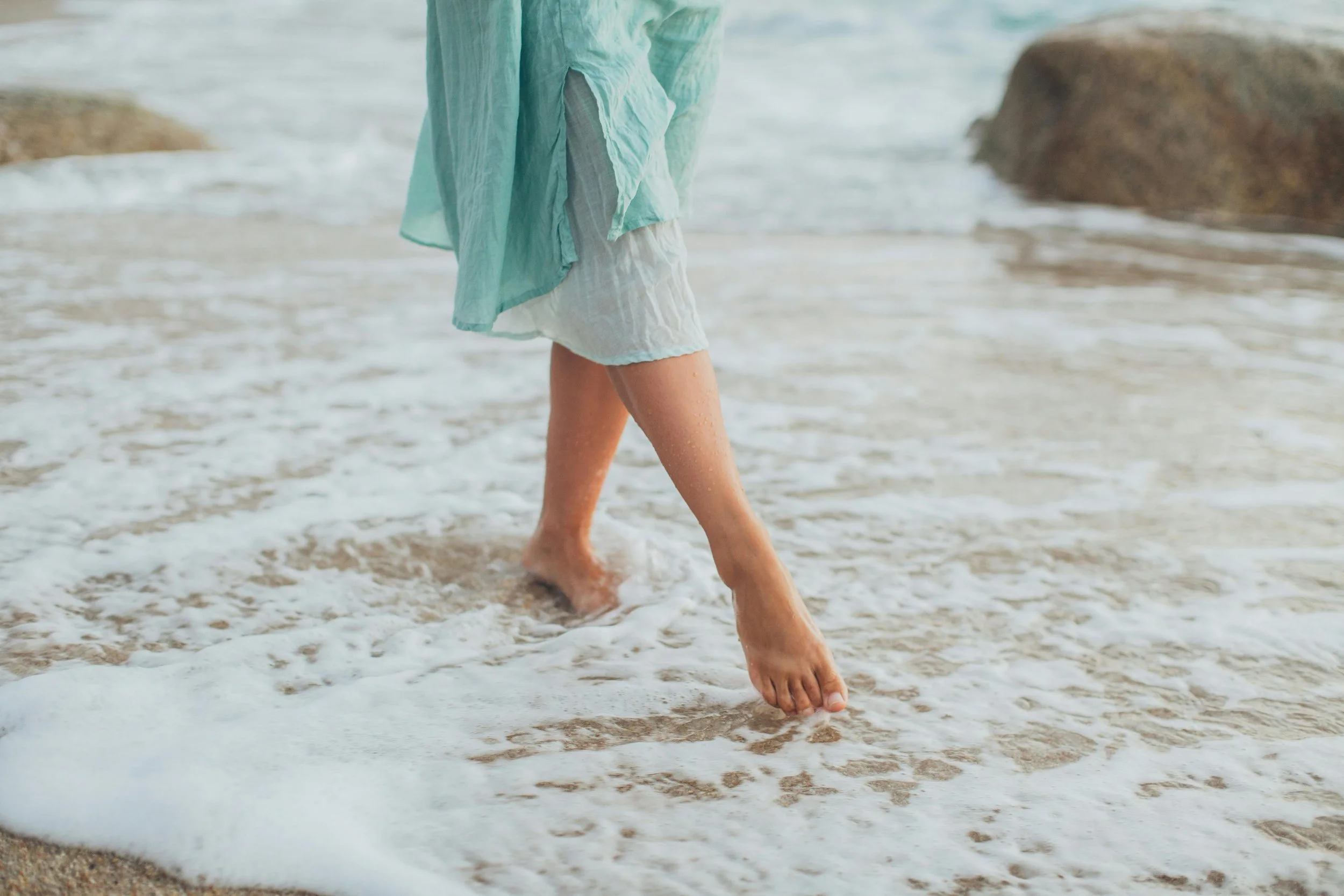 A person walking barefoot on the sandy beach with ocean waves washing over their feet, wearing a light green top and white shorts, with rocks in the background.