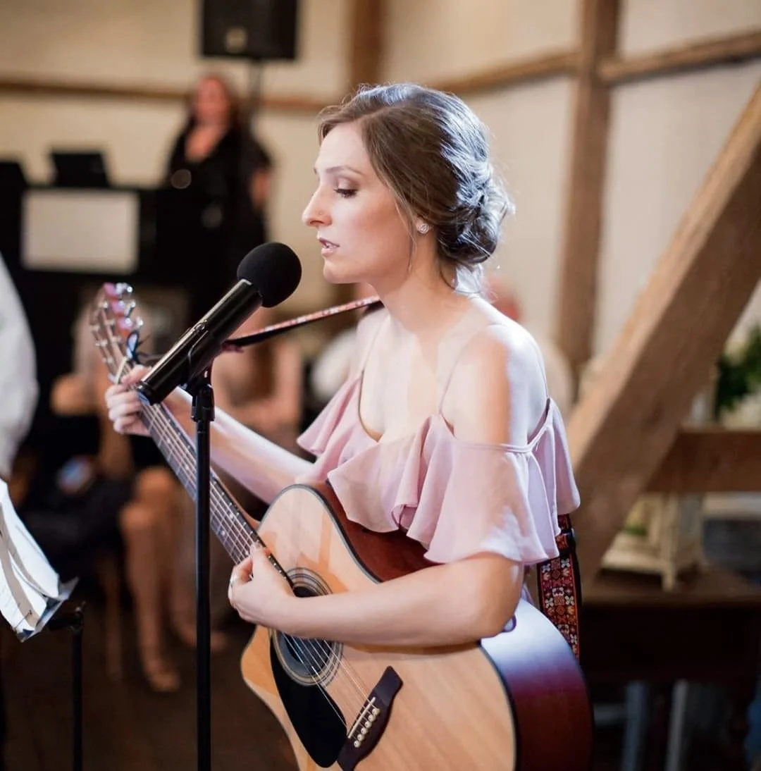 A woman with brown hair and a pink off-shoulder dress playing an acoustic guitar and singing into a microphone at an indoor event.