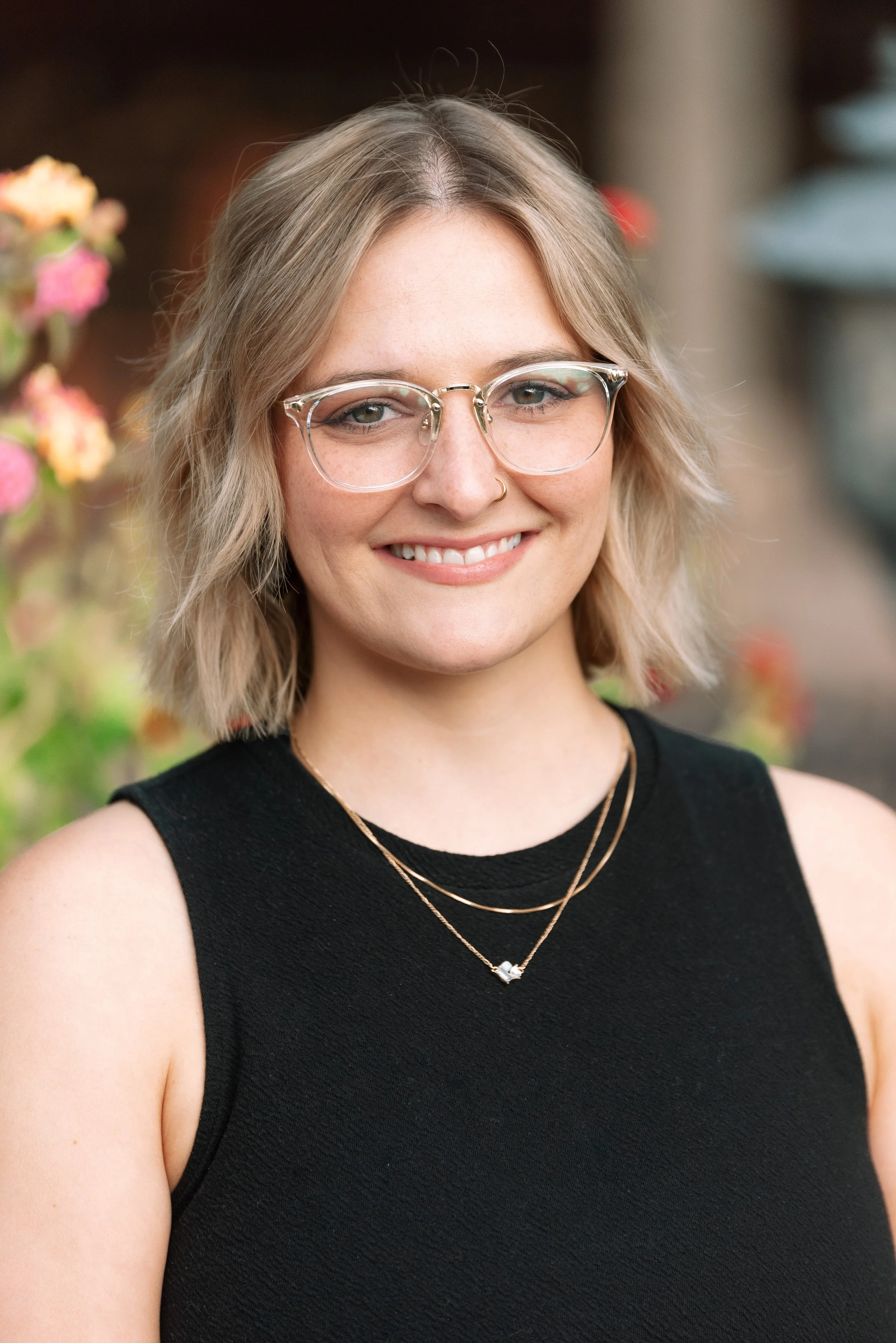 A young woman with shoulder-length blonde hair, wearing clear glasses, a nose ring, a black sleeveless top, and layered necklaces, smiling outdoors with pink and orange flowers in the background.